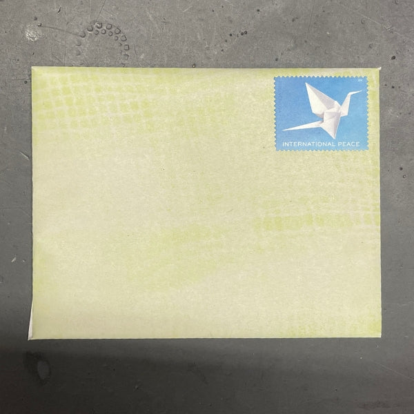 A square pale yellow envelope or card with a subtle grid pattern and a blue postage stamp featuring a white origami crane and the text 'INTERNATIONAL PEACE' in the top right corner, placed on a dark gray textured surface.